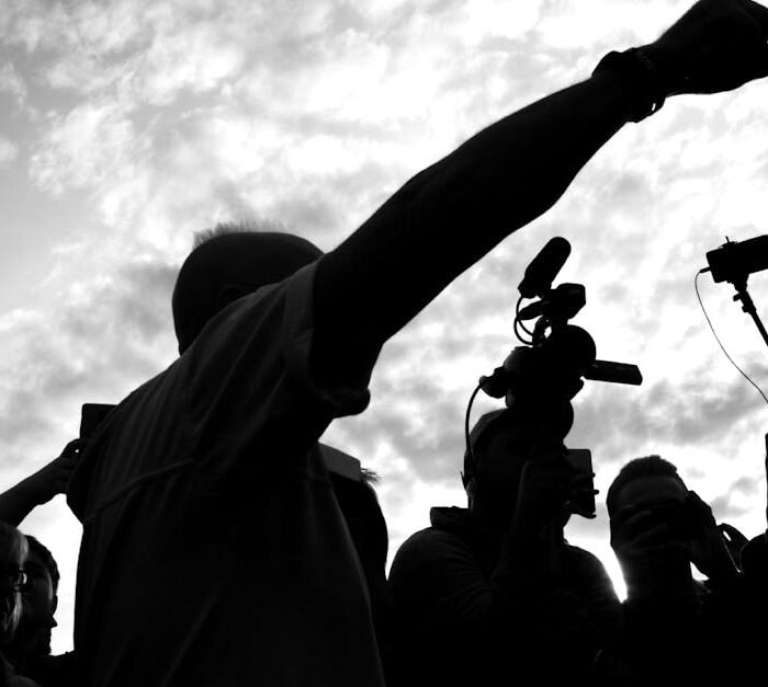 A dramatic silhouette of a media crew with cameras capturing a pointing individual against a cloudy sky.